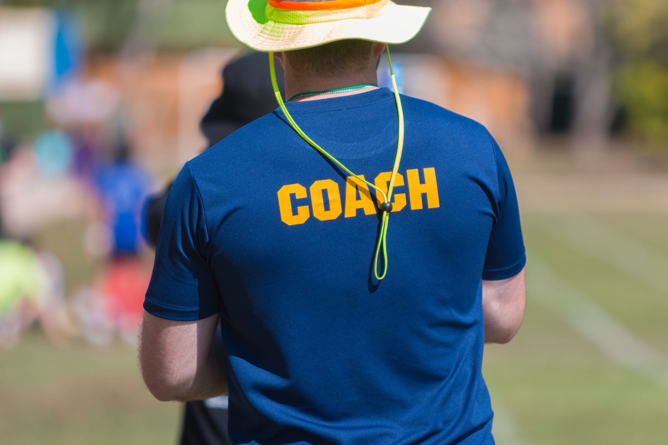 back of a coach's blue color shirt with the word Coach in yellow color written on, blurred outdoor sport field background
