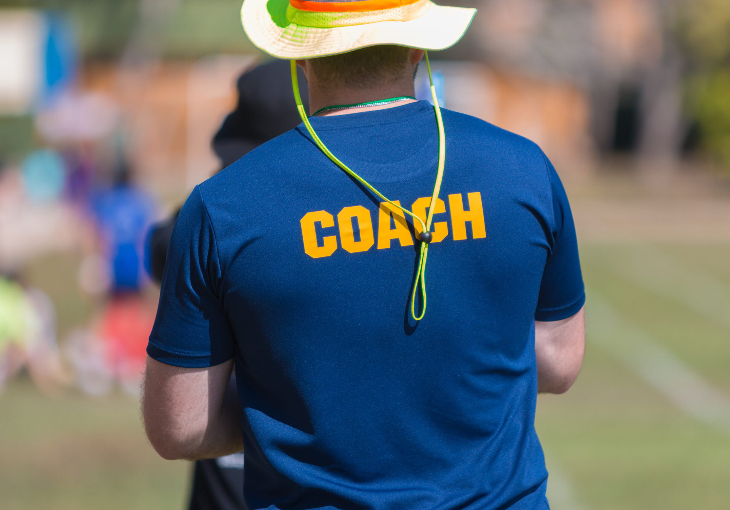 back of a coach's blue color shirt with the word Coach in yellow color written on, blurred outdoor sport field background